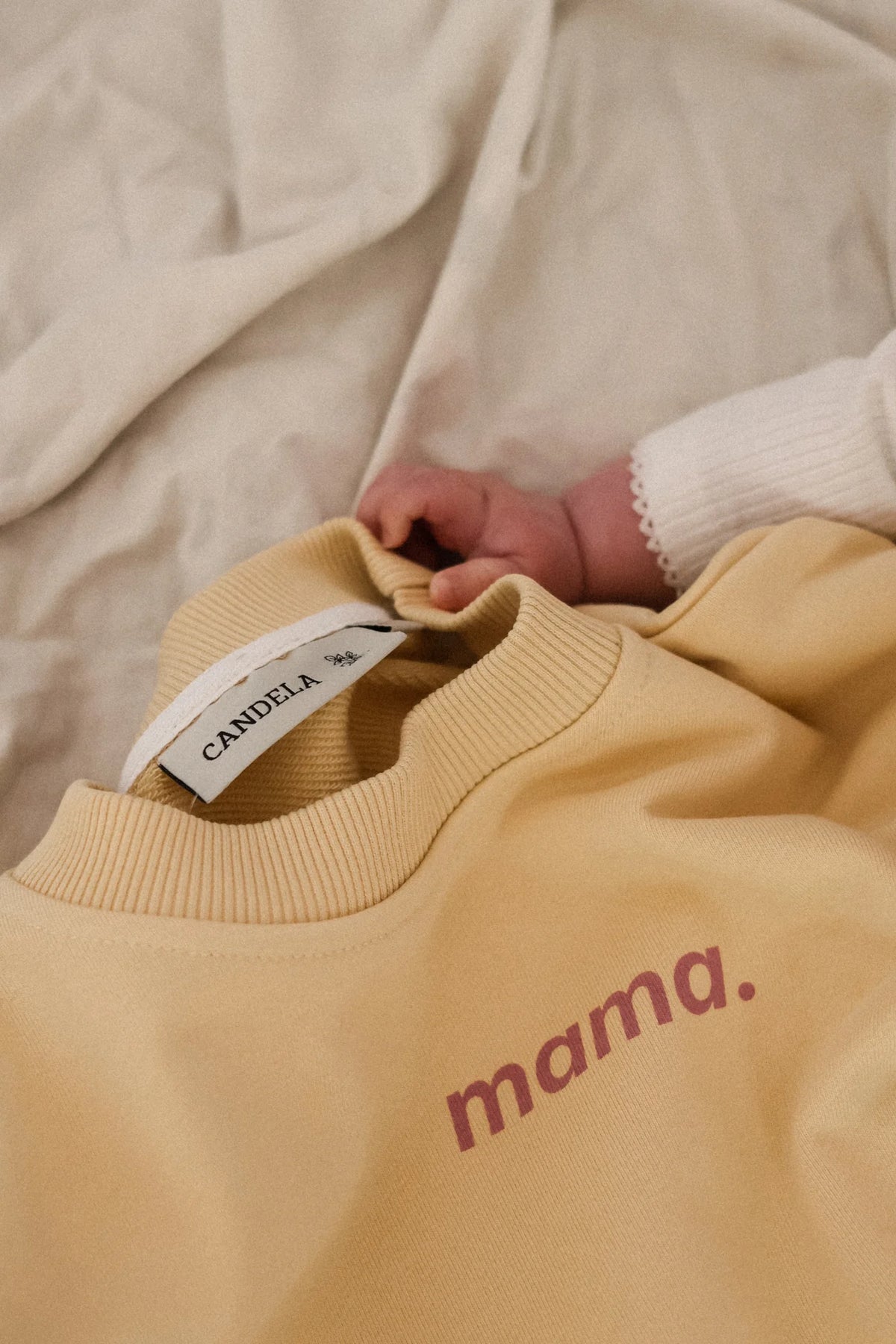 Zbliżenie na napis mama. i metkę CANDELA na bananowej bluzie Melanie, obok rączka niemowlęcia