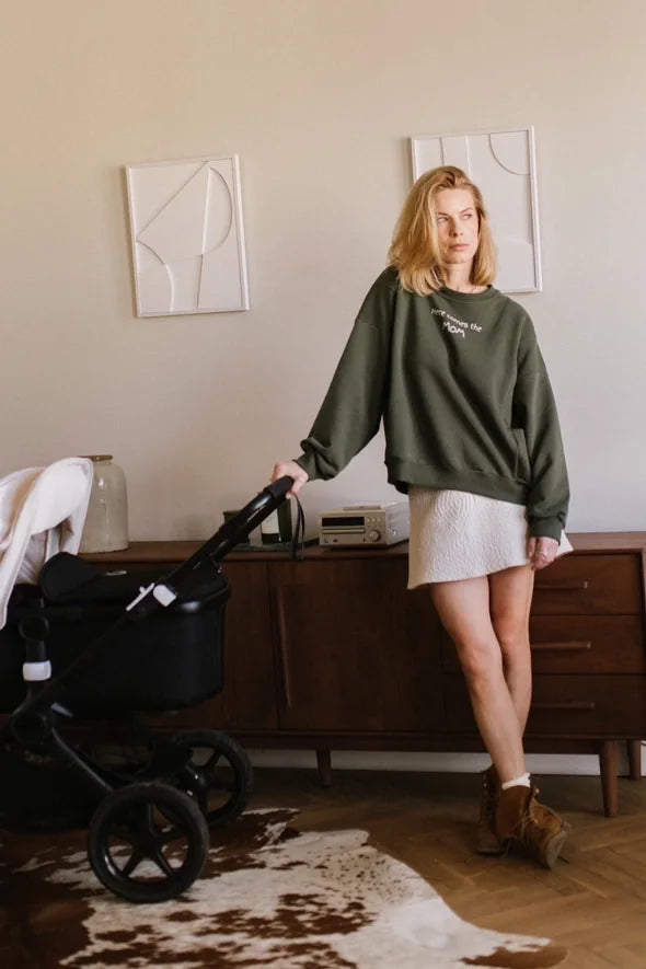 Kobieta w khaki bluzie oversize Melanie Here comes the MOM i jasnej spódnicy stoi przy dziecięcym wózku
