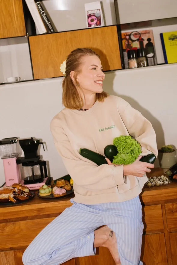 Kobieta w beżowej bluzie Melanie z zielonym nadrukiem Eat locally trzyma naręcze świeżych warzyw