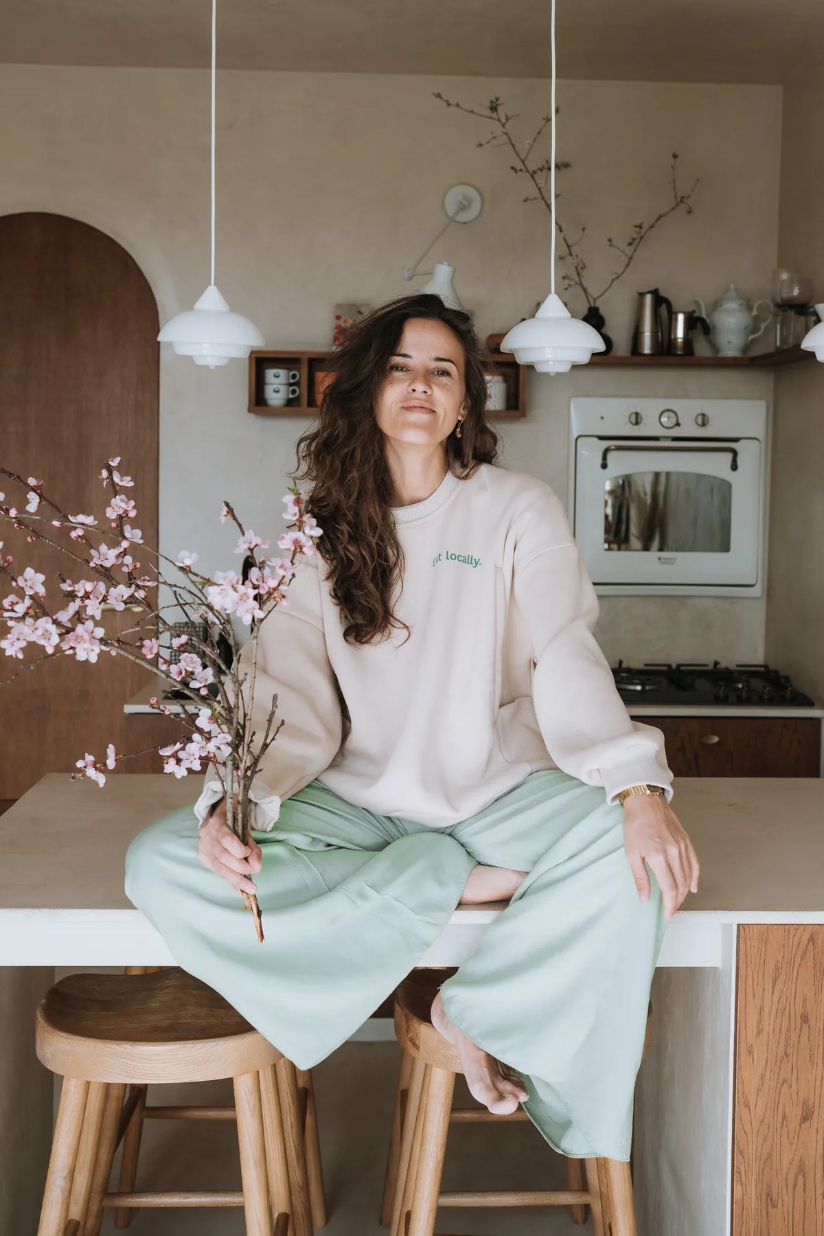 Kobieta w beżowej bluzie oversize Melanie Eat Locally siedzi na kuchennym blacie z gałązkami kwitnącej wiśni