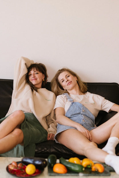 Dwie przyjaciółki odpoczywają na kanapie w beżowej bluzie oversize i t-shircie z kolekcji Eat Locally