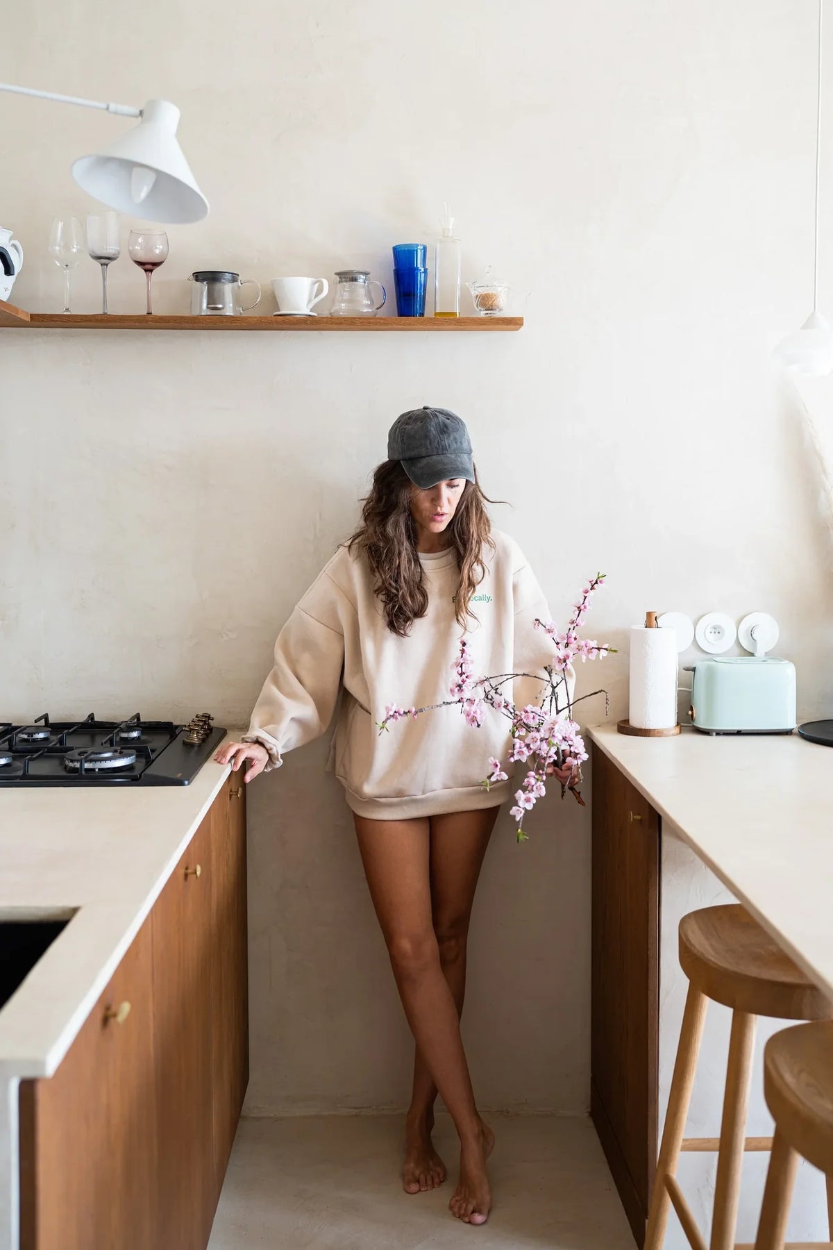 Kobieta w beżowej bluzie oversize Melanie Eat Locally i czapce z daszkiem stoi w kuchni z gałązkami kwitnącej wiśni