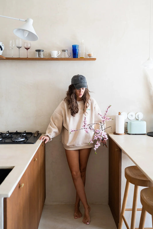 Kobieta w beżowej bluzie oversize Melanie Eat Locally i czapce z daszkiem stoi w kuchni z gałązkami kwitnącej wiśni