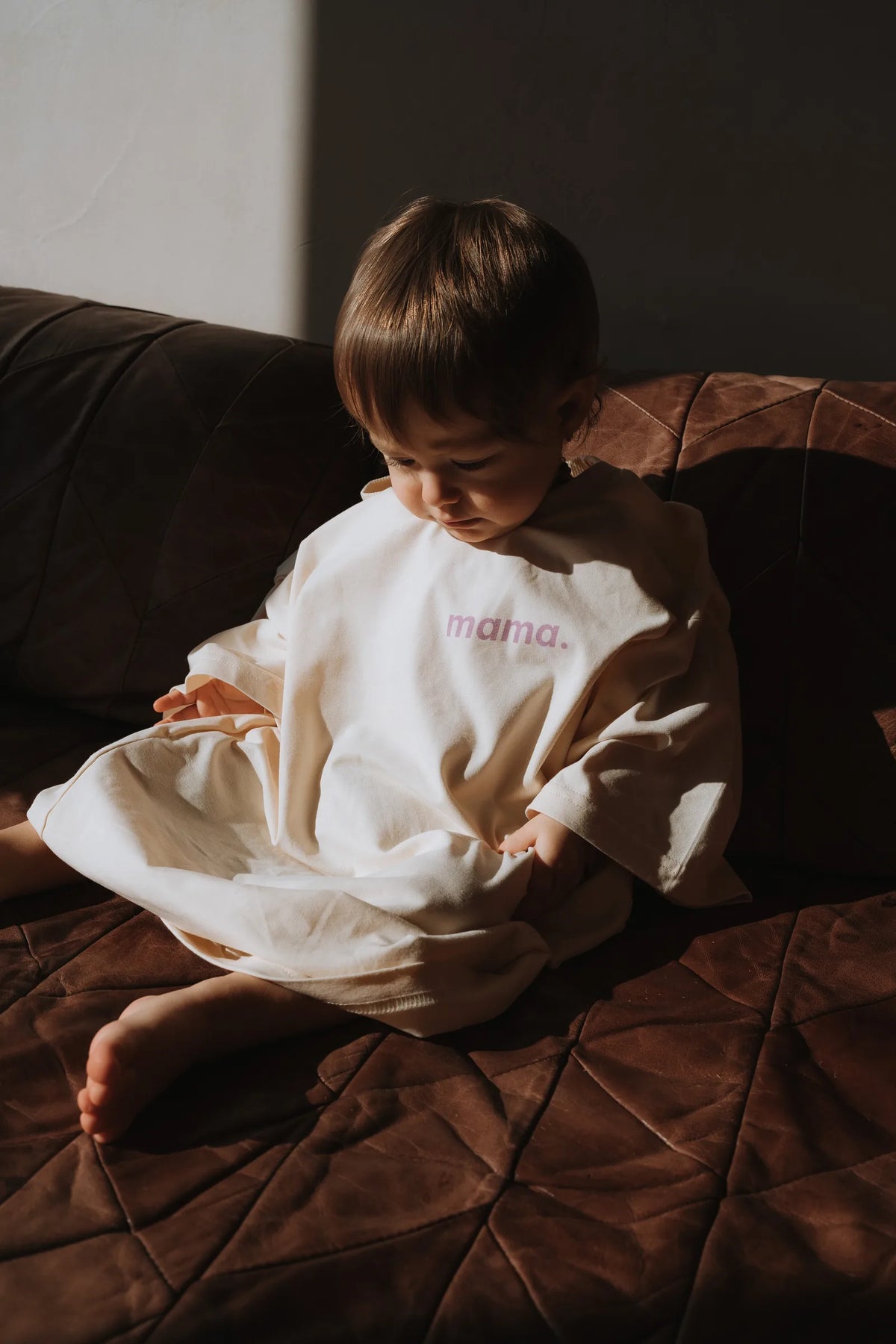 Maluch w za dużej kremowej koszulce z napisem mama. siedzący na brązowej skórzanej kanapie