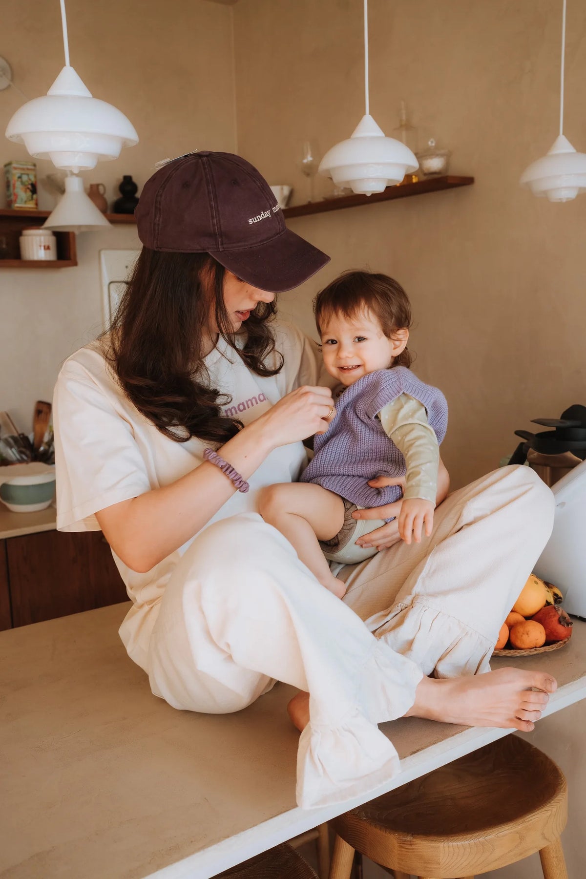 Mama w kremowym T-shircie do karmienia mama. i bordowej czapce trzyma uśmiechnięte dziecko w kuchni