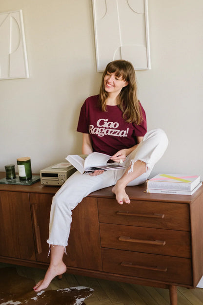 Uśmiechnięta kobieta w winnym t-shircie damskim Helen z napisem Ciao Ragazza siedzi na komodzie z książką