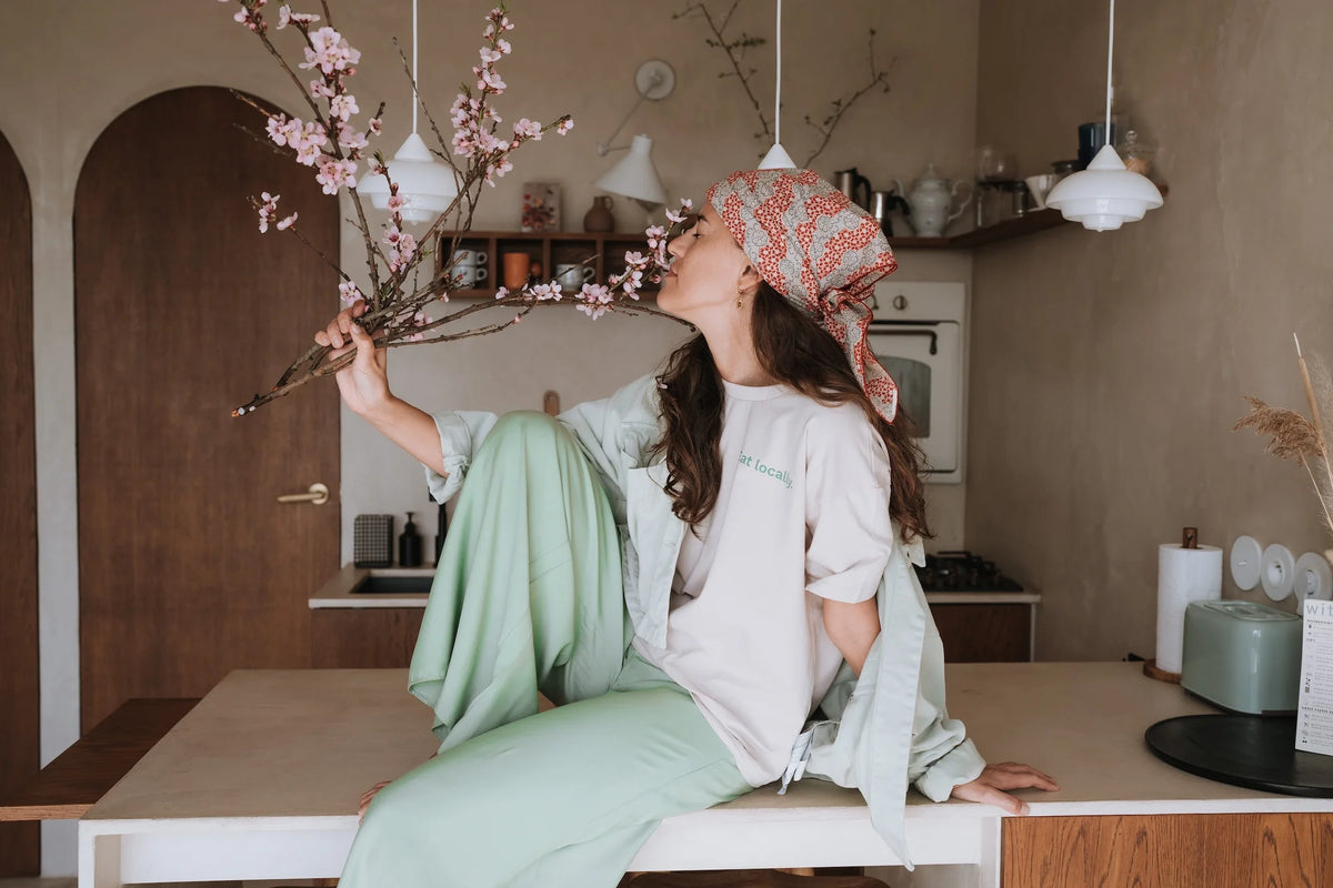 Kobieta w beżowym t-shircie Helen z zielonym napisem Eat locally i chustce na głowie wącha gałązki kwitnącej wiśni w kuchni