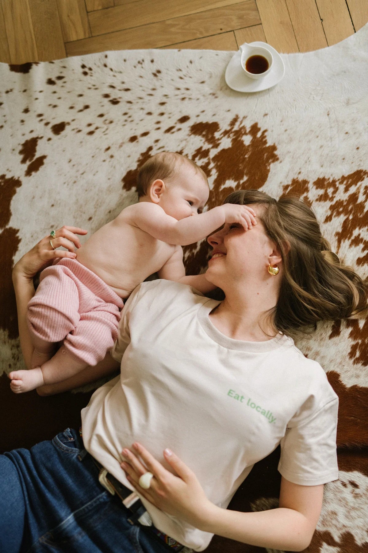 Uśmiechnięta mama w beżowym t-shircie Helen Eat Locally leży na dywanie i bawi się z niemowlęciem