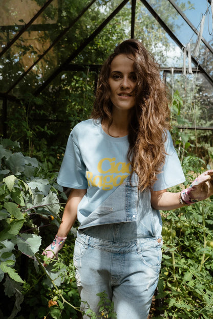Kobieta w niebieskim t-shircie oversize z żółtym napisem Ciao Ragazza i jeansowych ogrodniczkach w szklarni