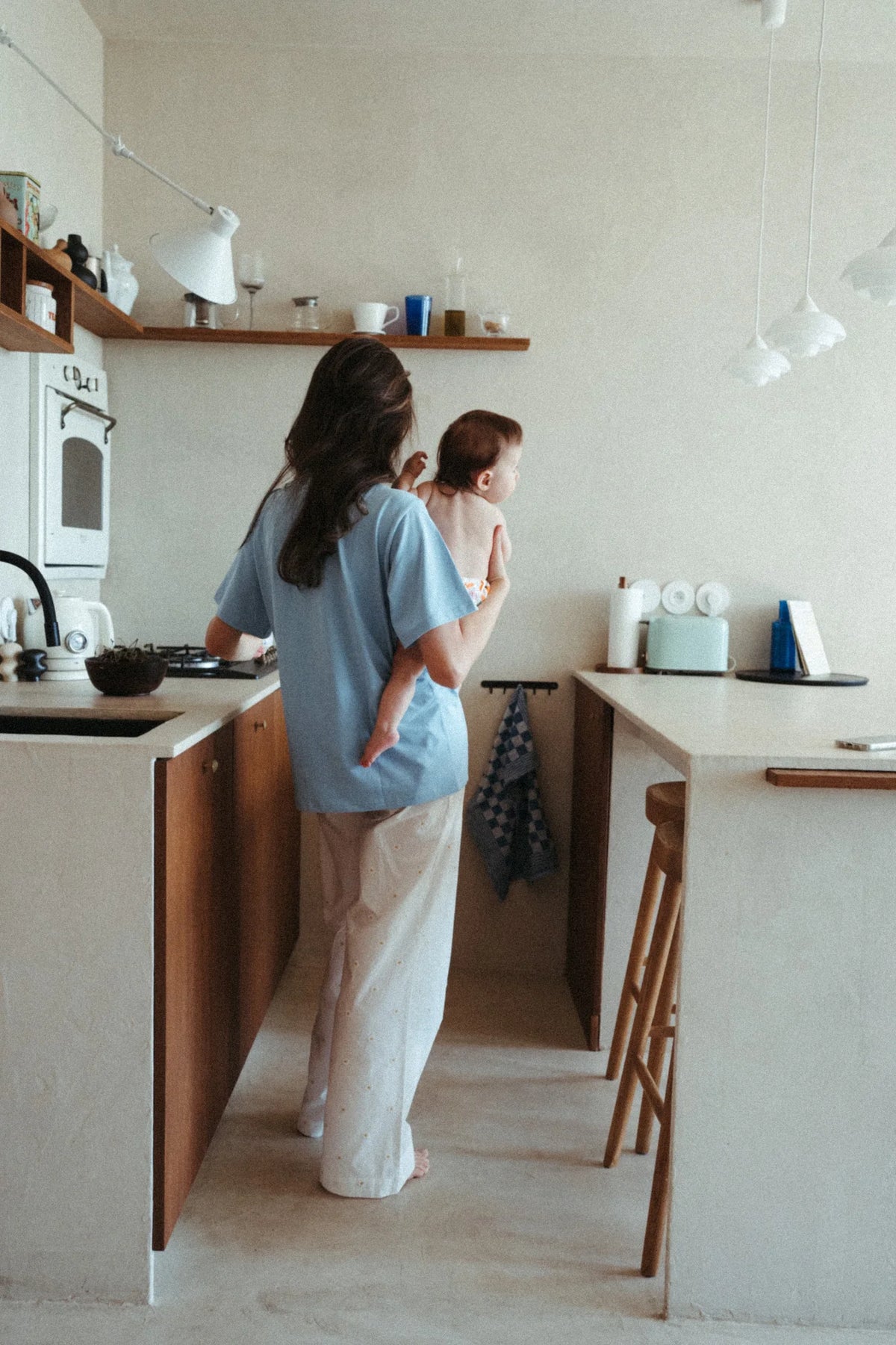 Mama w niebieskim t-shircie oversize Ciao Ragazza trzyma niemowlę na rękach w kuchni, widok z tyłu