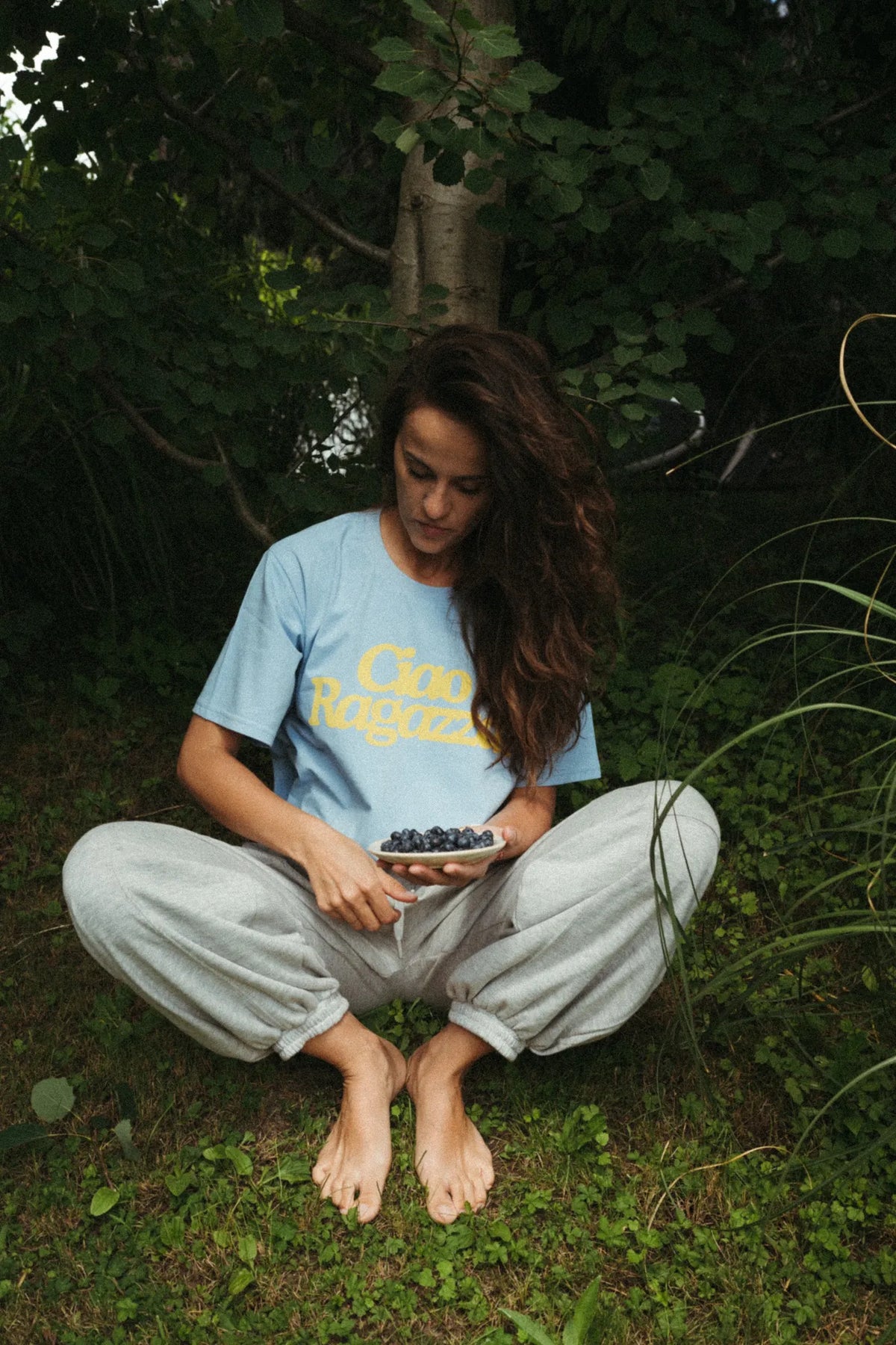 Kobieta w niebieskim t-shircie z żółtym napisem Ciao Ragazza siedzi na trawie z miseczką jagód