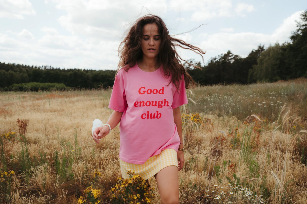 Kobieta w różowym t-shircie oversize z czerwonym napisem Good enough club i żółtej spódnicy w kratkę idzie przez łąkę