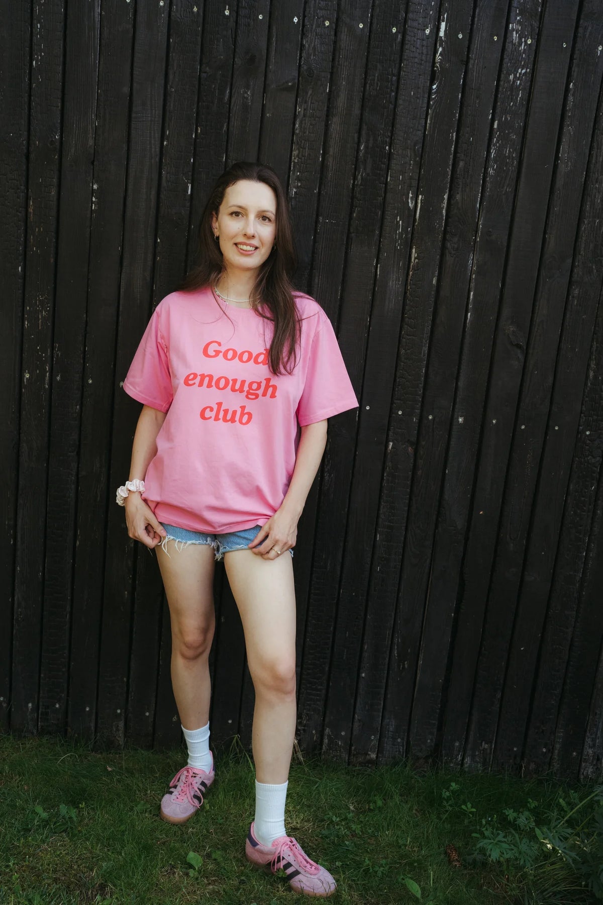Uśmiechnięta kobieta w różowym t-shircie damskim oversize Good enough club i jeansowych szortach przy drewnianej ścianie