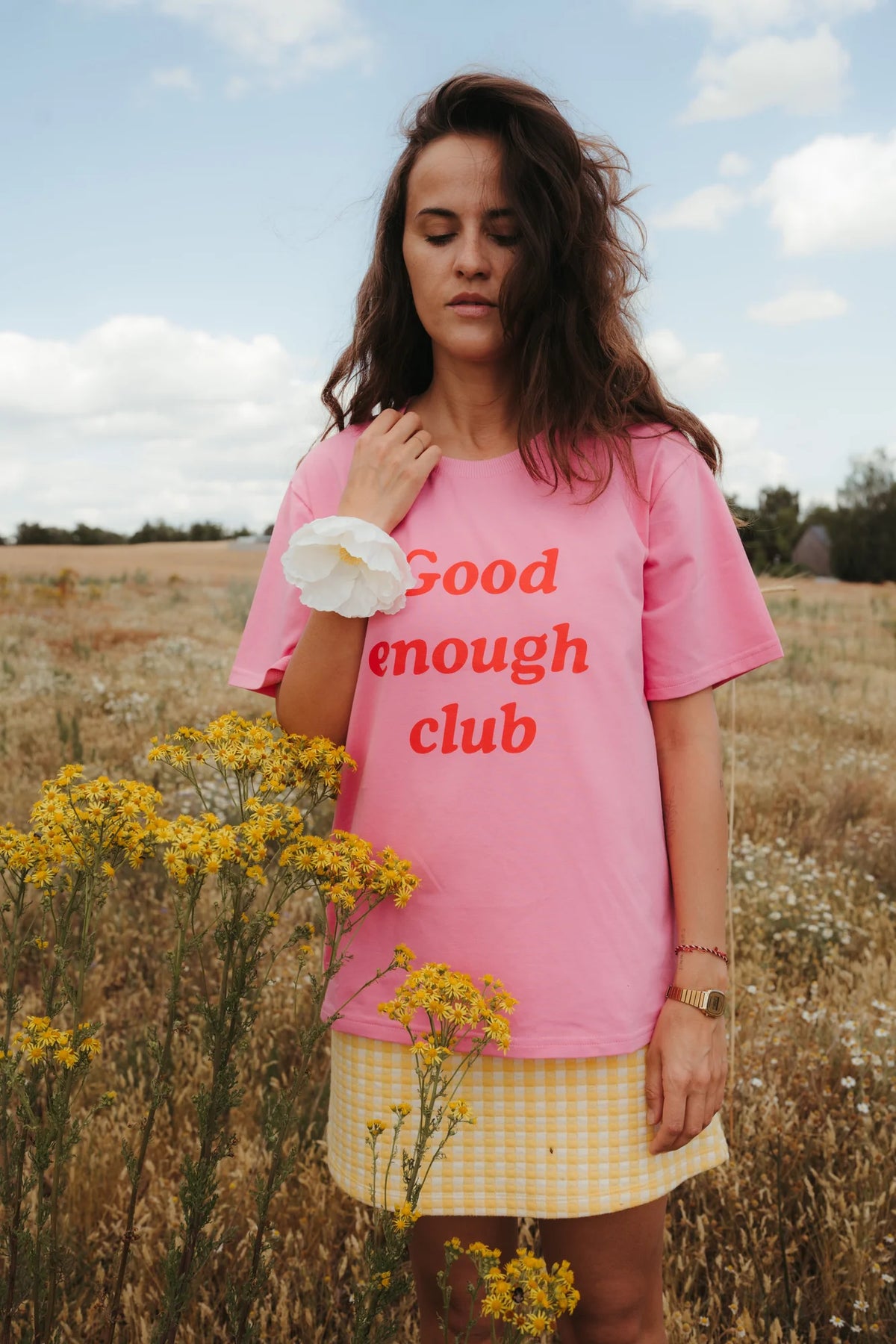 Kobieta w różowym t-shircie oversize Good enough club stoi na łące wśród żółtych kwiatów