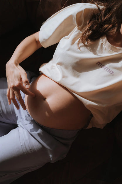 Kobieta w ciąży w kremowym t-shircie oversize mama. i lawendowych spodniach odsłania brzuszek