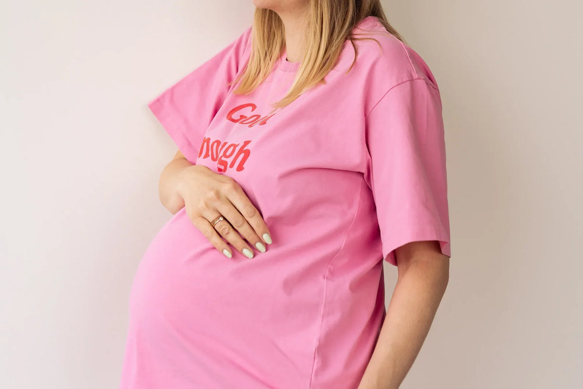 Kobieta w ciąży trzyma dłoń na brzuszku w różowym t-shircie do karmienia oversize Good enough club